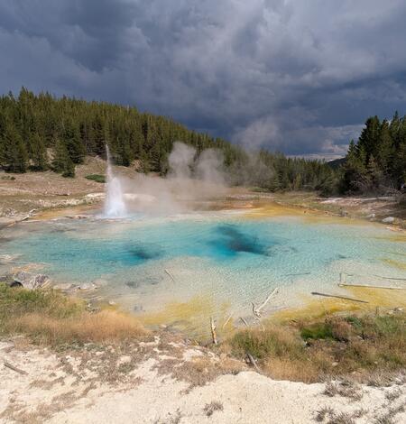 blue-water hot spring with a geyser to one side, forest in background, and storm clouds in the distance