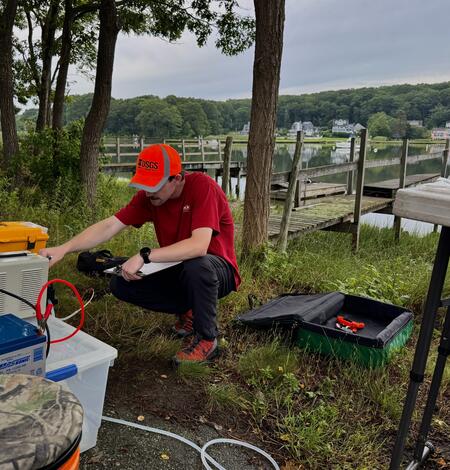 A man wearing a red shirt and orange hat crouches down in front of a peristaltic pump in front of a dock.