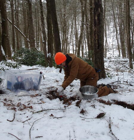 A person with a brown outfit and orange hat takes a soil sample in the woods with snow on the ground.