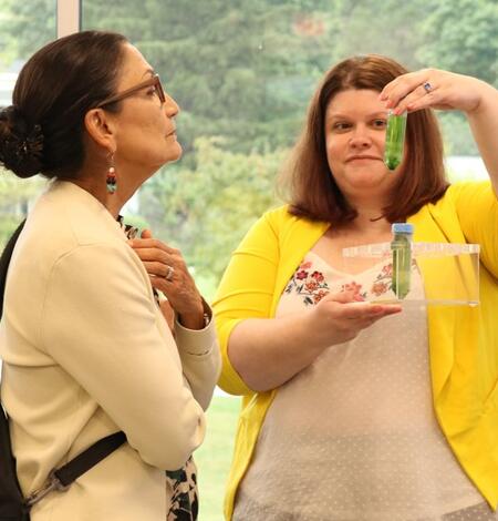 A woman holds a test tube with green water up to show another woman peering at it through her glasses
