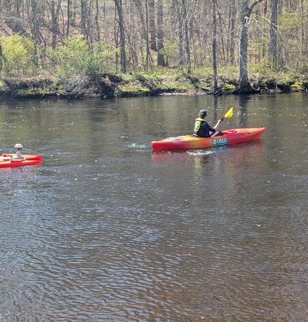 A woman kayaks upstream while towing an orange ADCP behind the kayak. 
