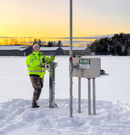 Allison standing in snow next at groundwater well site with three large buildings in the background