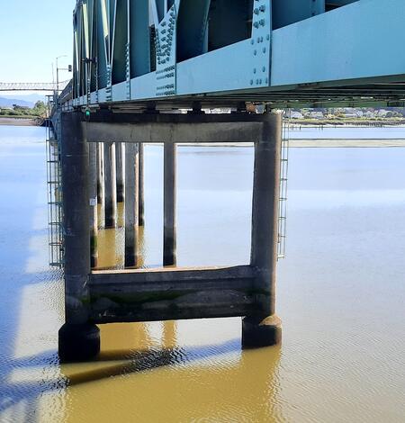 High bridge stretches across a large expanse of water. Grey bridge pillars hold up the blue-green steel bridge