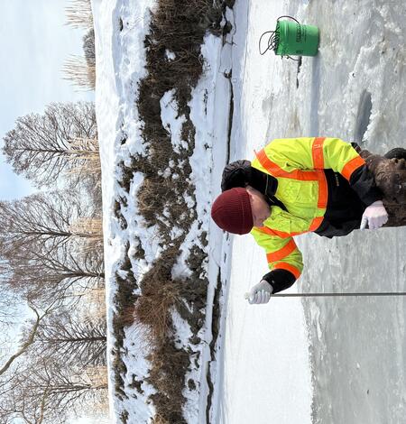 Hydrologic technician in bright yellow coat collects water sample through hole in ice on frozen river