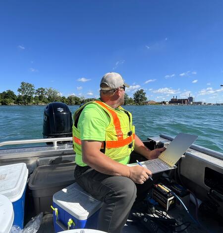 Hydrologic technician on a boat, holding a laptop looks over a blue river