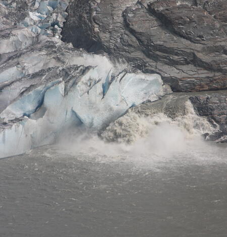 White, frothy water pours from the bottom of a blue glacier, marring the surface of a calm grey lake.