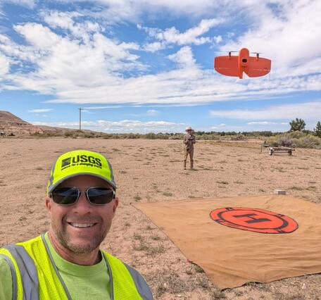 two people stand in a dirt landscape with a UAS in the air and helipad on the ground