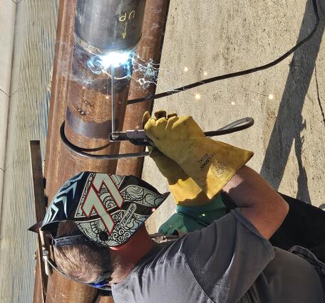 A welder in a helmet and heavy gloves welds a joint between two sections of steel well casing pipe on the ground beside a can