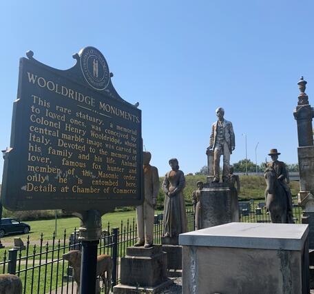 Historical marker installed beside weathered human-figure statues, all enclosed by an iron fence.