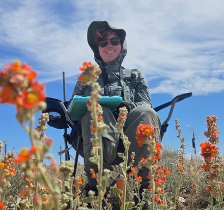 A person sitting in a chair with a clipboard smiles, there are orange globemallow flowers in the foreground and a cloudy blue sky in the background