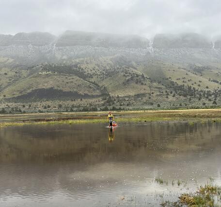 A female in safety gear pulling equipment through the water looking small amidst the stunning background of misty hills