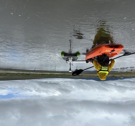 back view of a kayaker paddling to push measurement equipment pontoon across calm waters on a cloudy day