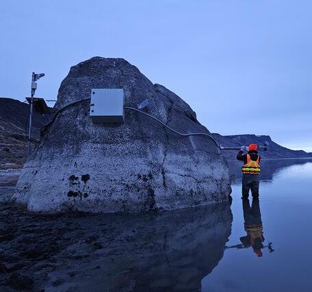 technician in safety gear stands near a large rock amidst the shallow lake waters reflecting a steel-blue early morning sky