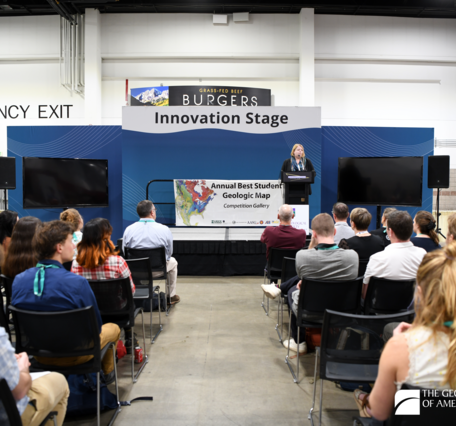 A person standing at a podium on a stage addresses a seated crowd. Behind presenter are two banners reading “Innovation Stage” and “Annual Best Student Geologic Map Competition Gallery.”