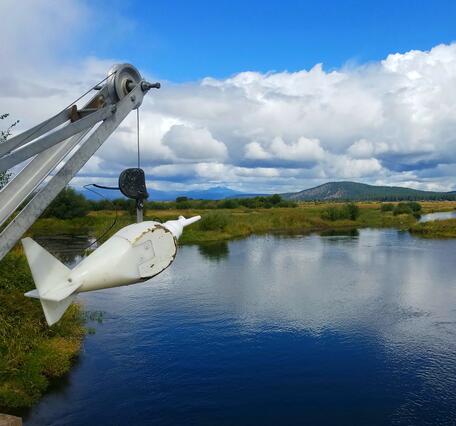 partly cloudy day. A white torpedo shaped sediment sampler hangs from a crane over a bridge with blue water below