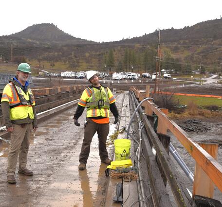 Two scientists in bright colored safety gear and hard hats stand on a bridge above the river.