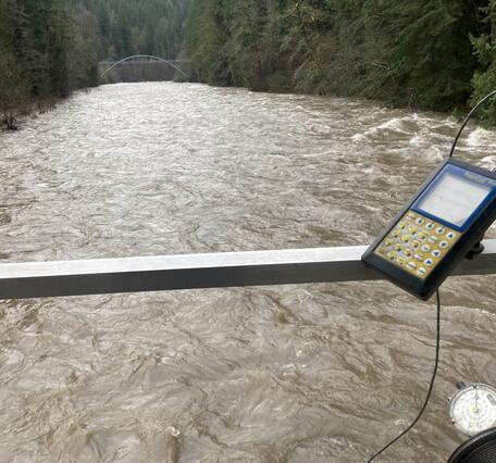 A swollen brown river fills the view. A flow calculator interface is fastened to aluminum rail of cable car.