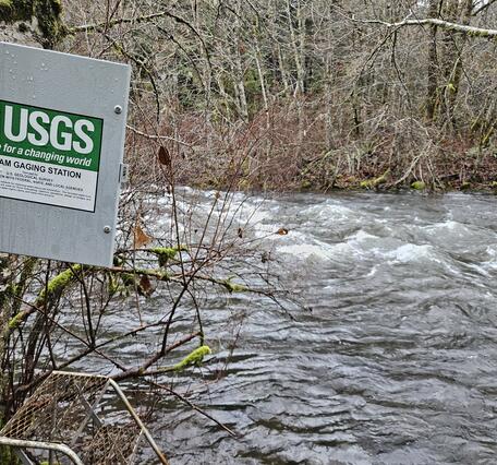 square metal enclosure door with USGS signage swings open by a rushing river