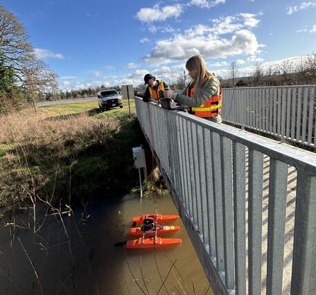 two people standing on a foot bridge look over metal railing at measurement equipment in the creek