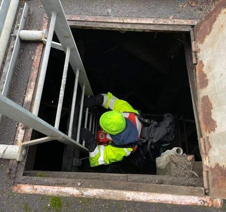 a person in bright safety gear descends a metal ladder through a square hole down into darkness