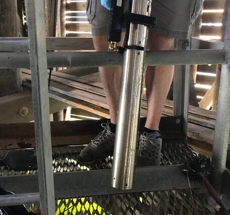 Male standing on metal platform holds cylindrical nitrate sensor. Green lit water shines from river beneath platform.