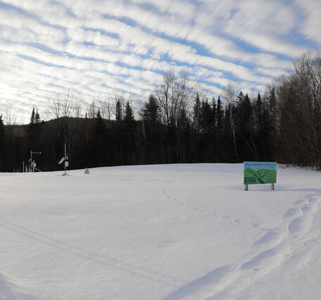 A snowy field with a blue and green sign on it and some meteorological equipment.
