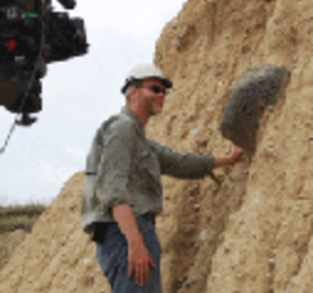 A geologist stands at an outcrop containing a small boulder deposited by a glacier with a video camera in the foreground. 
