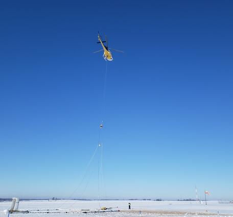 Helicopter with large hoop hanging below, technician on snowy ground for scale