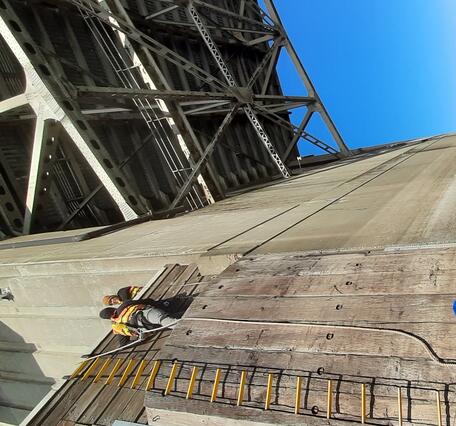two people are high up on a bridge pier ledge while another crew assists from a boat parked close to the bridge pier. 