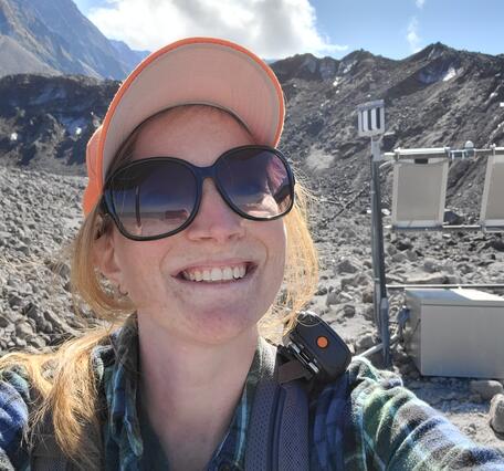 A young woman in dark sunglasses and an orange hat crouches next to a scientific sensor on a rocky slope. She has blonde hair and fair skin.