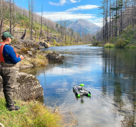 Male scientist operates a remote-controlled device from the riverbank in a burned forest landscape.