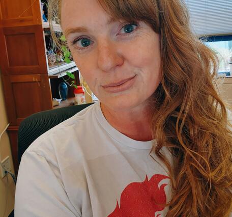 A woman with long hair leans toward the camera with a bookcase and window in the background.