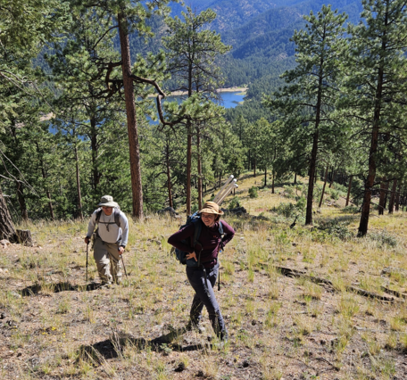 two people walking uphill through a pine forest, with a lake and mountains in the background