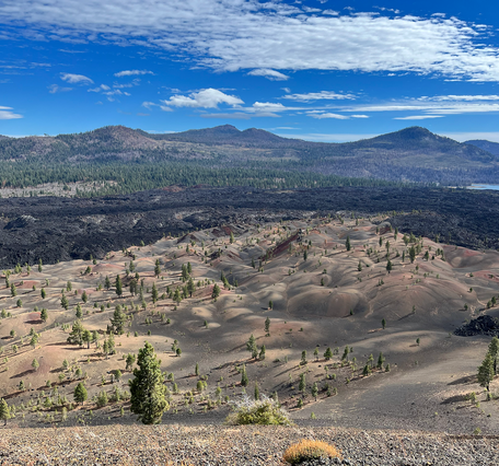 Taken from a high vantage point, this photo shows a landscape of rolling, barren hills stained in reds, tans, and blacks merging into a field of jagged black lava flows. In the distance, mountains rise above a pine-forest and bright blue lake.