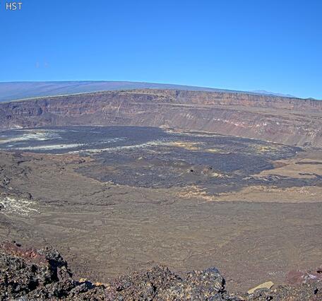 Desolate volcanic crater of gray/brown rick under a blue sky with a sloping mountain in the background