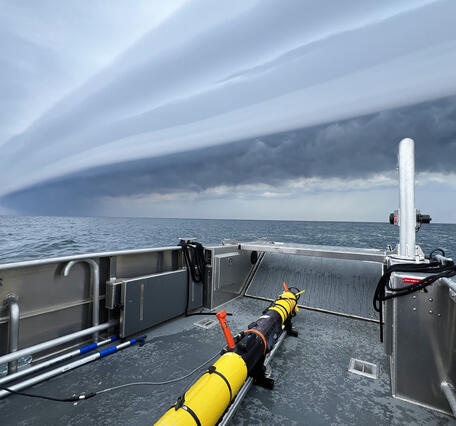 A long yellow autonomous vehicle sits on the deck of a research vessel on a stormy day.