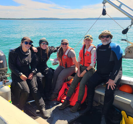 Five researchers posing on the deck of a small vessel on Lake Michigan. An engine and net sampling gear are present.