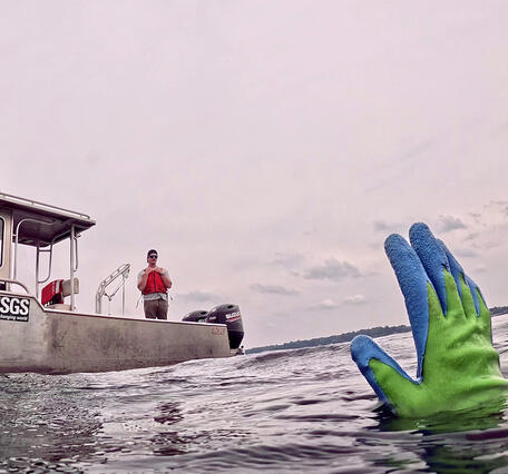 The gloved hand of a diver emerges from a lake and signals researchers on a small vessel, R/V Sander, in the background