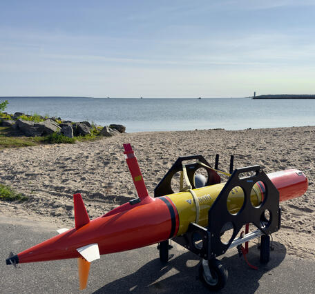 An orange Underwater Autonomous Vehicle in a transport cradle on a beach with a lake in the background.