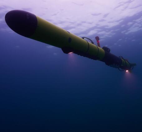 A view underwater in Lake Michigan looking up at a yellow Autonomous Underwater Vehicle with two lights shining down.