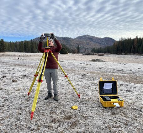 Setting up Global Navigation Satellite System base station on a tripod in a forest clearing with mountains in the background