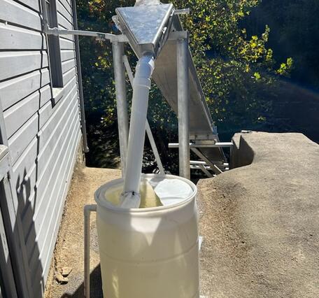 A white barrel sits beside a building, with PVC pipes feeding into it from a metal chute above.
