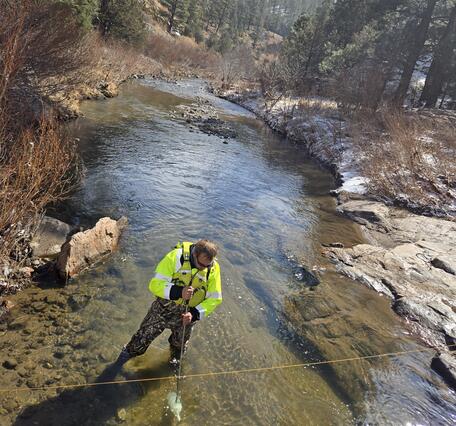 A hydrologist stands in a low-flowing creek and is holding a sampling bottle under water