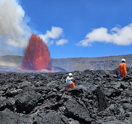 Color photograph of scientists on crater floor with lava fountain visible in the background