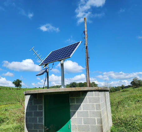 A small concrete block wellhouse with two solar panels and a satellite antenna on the roof.