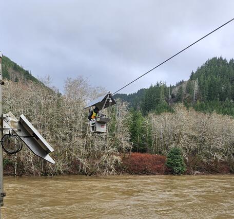 A man suspended above murky rushing river waters measures flow from an aluminum cable car