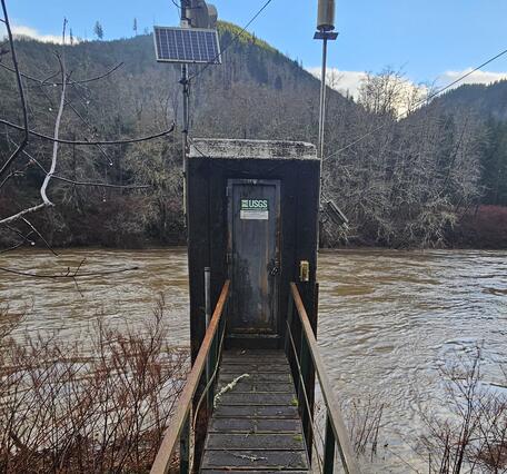 Square concrete structure built alongside the riverbank has solar power & antennas on top and wooden walkway leading to door