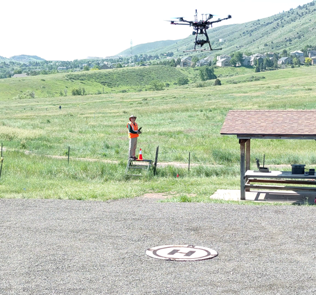 Pilots in a grassy field by a picnic shelter containing their instruments stand below a hovering drone