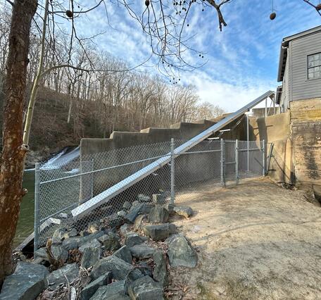 A concrete dam structure along a wooded river with a metal chute and chain‑link fencing beside a gray building.