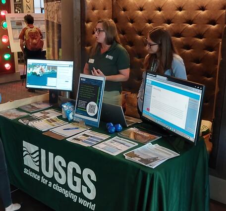 Two USGS scientists behind a booth filled with handouts and computer monitors talking to two visitors